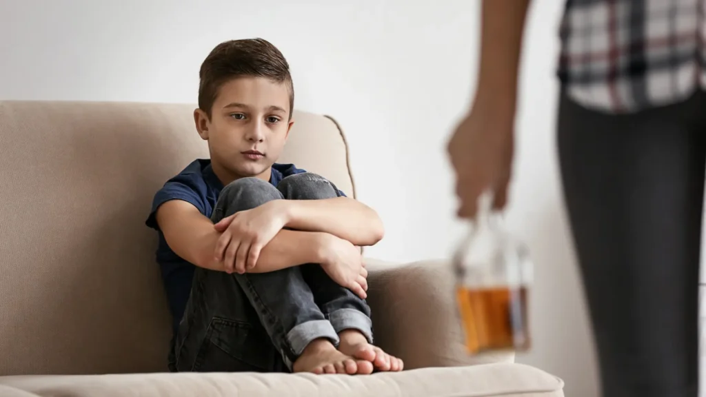Smutny chłopiec na kanapie, w tle dorosły z butelką alkoholu, syndrom DDA
